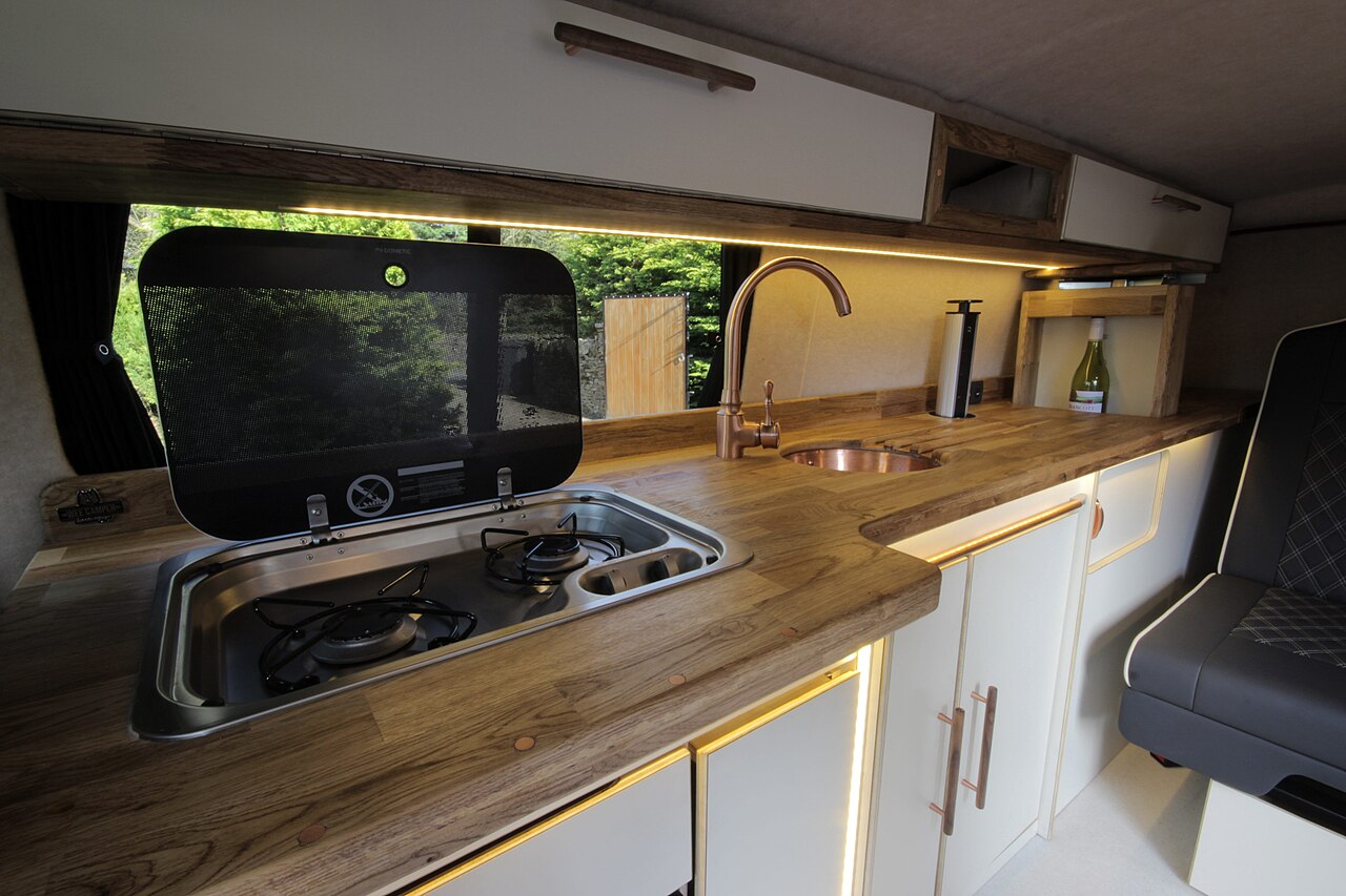 Ausgebauter Innenraum eines Campervans mit Küchenzeile, Gaskochfeld, Spüle, Holzarbeitsplatte und Oberschränken