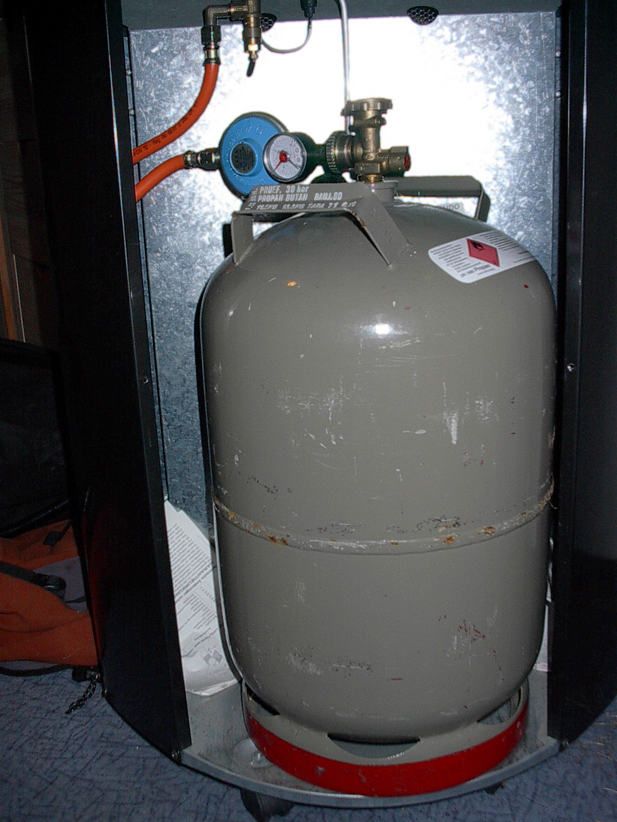 Graue Propan-Stahlflasche mit Druckregler, Manometer und orangefarbenem Gasschlauch in einem Schrank.