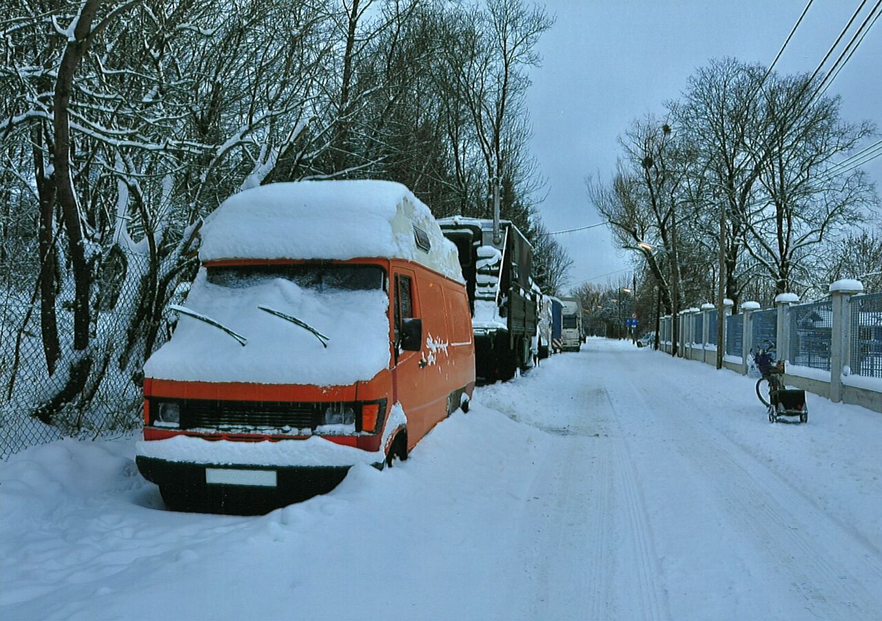 Eingeschneiter orangefarbener Campingbus am Straßenrand, Dach und Frontscheibe mit Schnee bedeckt.