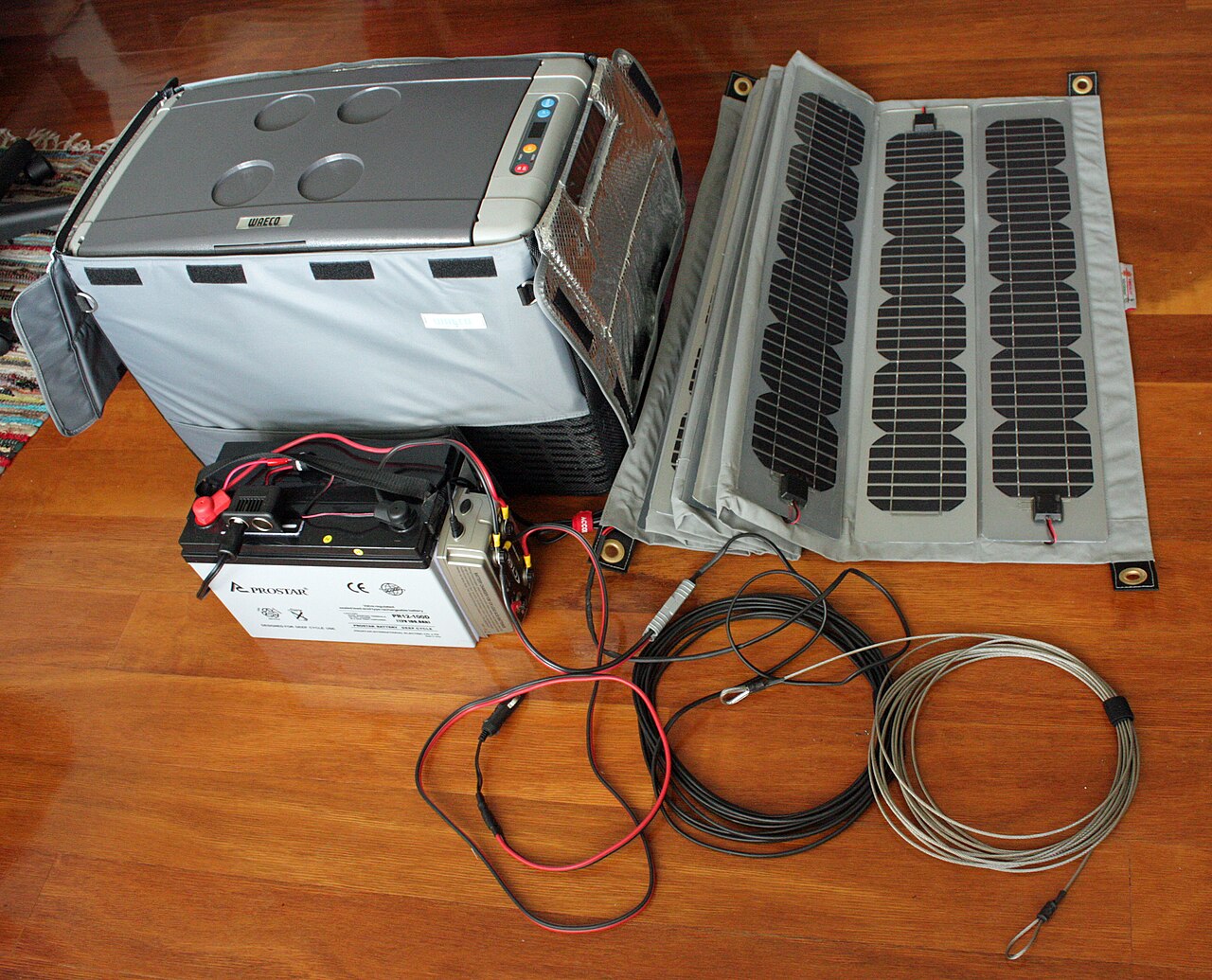 Ein faltbares, flexibles Solarpanel mit drei Modulstreifen auf einer Stoffmatte, daneben Zusatzbatterie, Kühlbox und aufgerollte Kabel auf einem Holzboden.